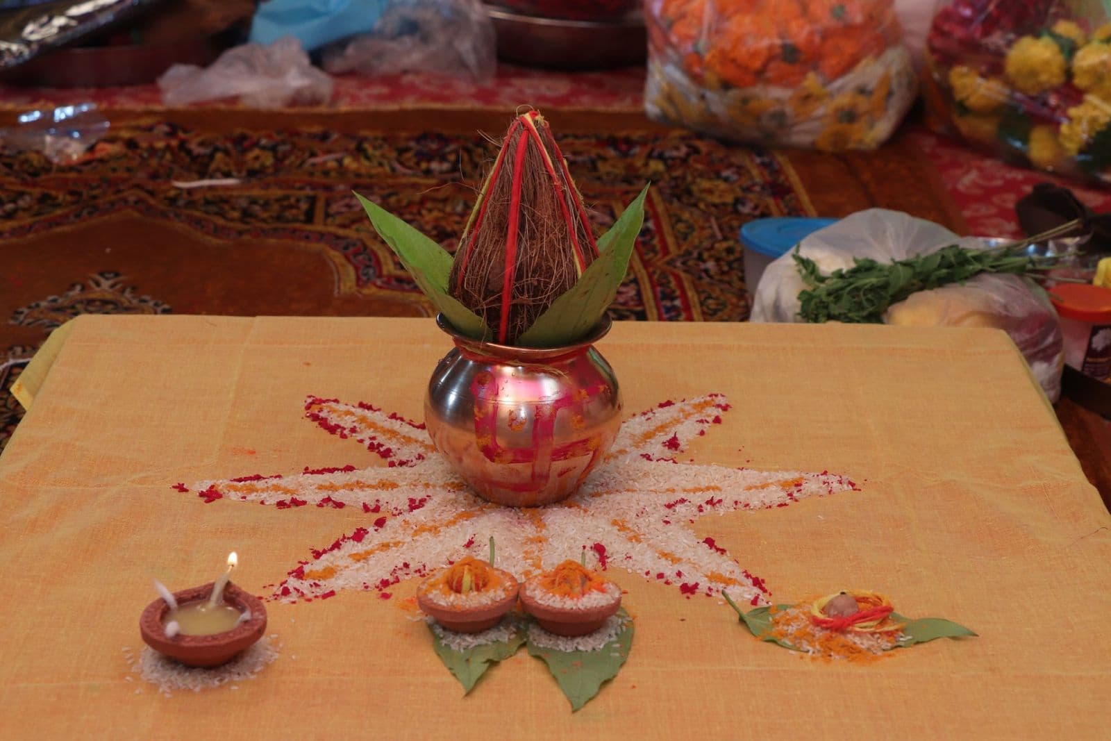 A copper kalash with mango leaves and a coconut on a flower rangoli, with small oil lamps, for an engagement ceremony